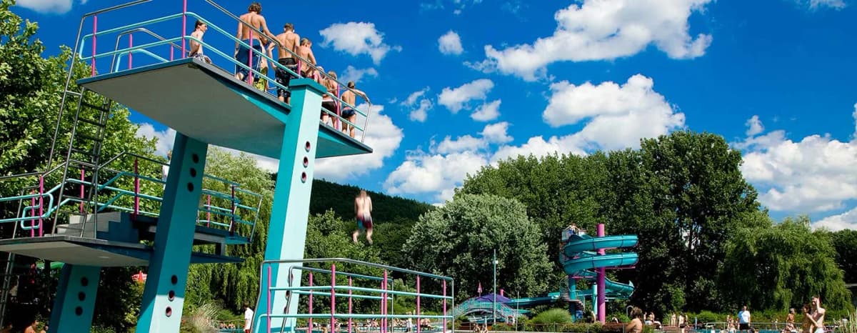 Sprungturm im Freibad mit Jugendlichen, einer springt gerade, im Hintergrund eine Wasserrutsche und Bäume