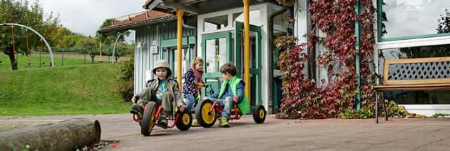 Kinder fahren mit Dreirädern auf dem Hof eines Kindergartens vor einem Gebäude mit rankendem Wein