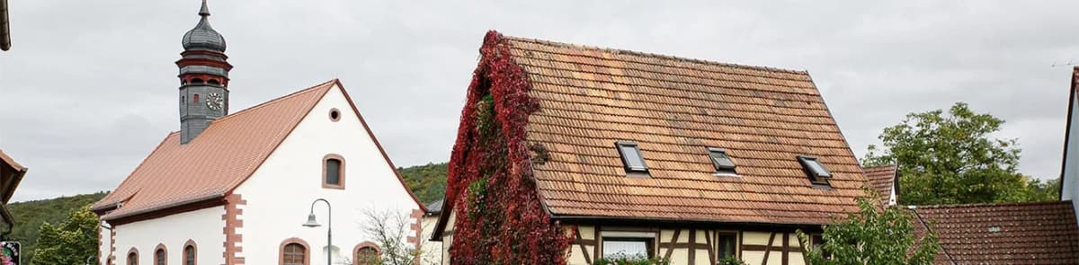 Kirchturm und Fachwerkhaus mit rot-braunem Ziegeldach, teils mit rotem Wildem Wein bewachsen