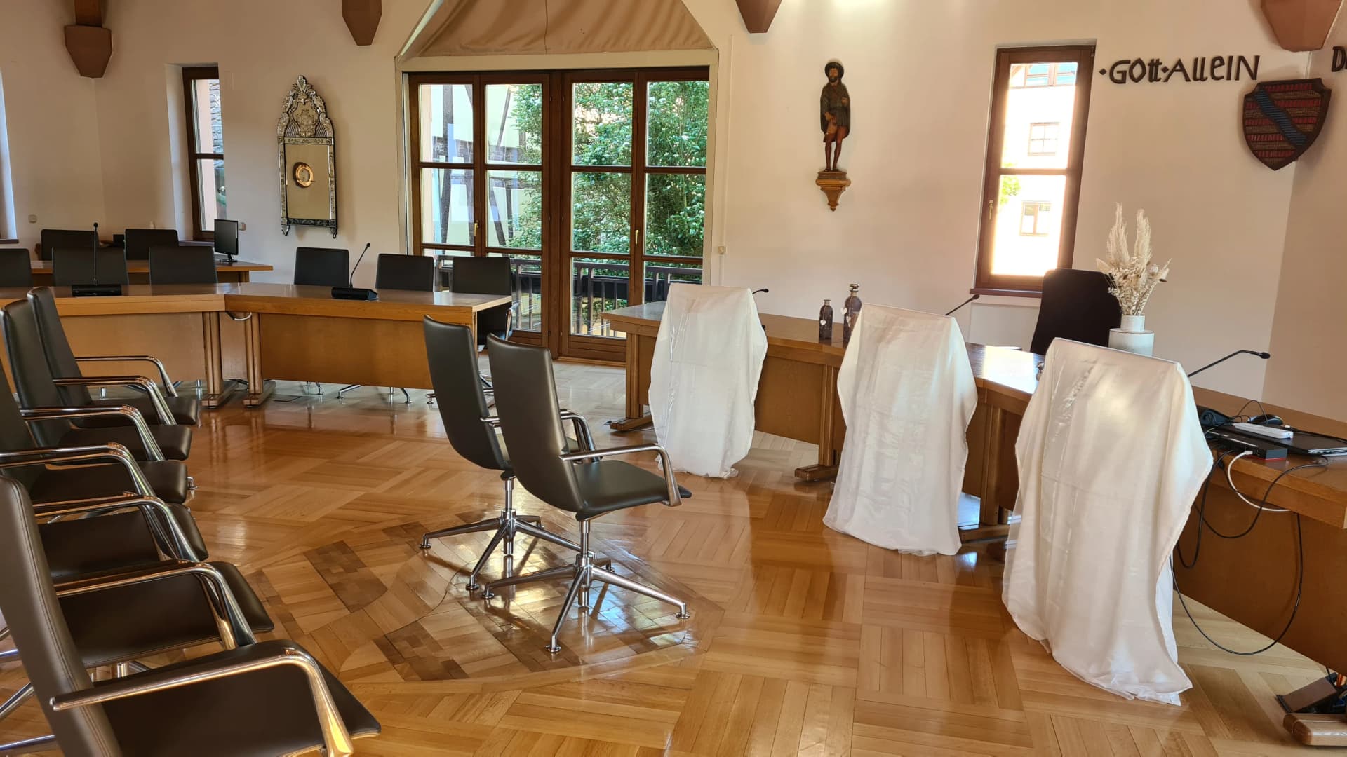 Sitzungssaal mit Stuhlreihen und Tischen, drei Stühle mit weißen Hussen bedeckt, an der Wand die Inschrift Gott Allein und eine Heiligenfigur
