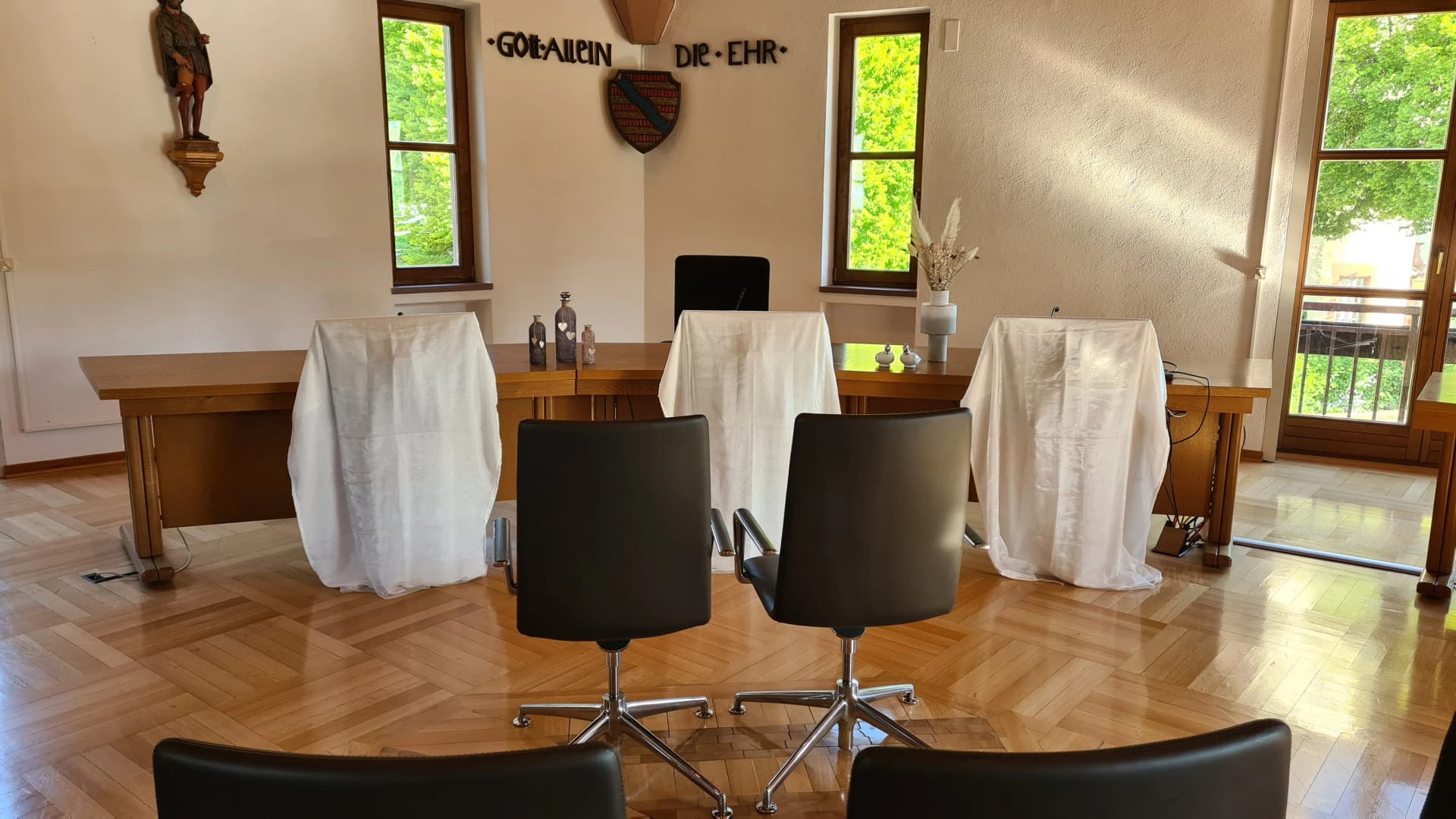 Sitzungssaal mit Rednerpulten unter weißen Tüchern, davor Stühle, an der Wand die Inschrift 'Gott allein die Ehr' mit Wappen und einer Heiligenfigur