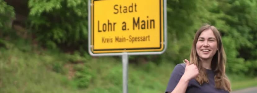 Frau mit erhobenem Daumen vor dem Ortsschild Stadt Lohr a. Main, Kreis Main-Spessart