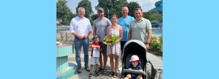 Gruppe von vier Männern, einer Frau mit Blumenstrauß, einem Jungen und einem Kleinkind im Kinderwagen posiert lächelnd im Freibad vor Bäumen und Rutsche.