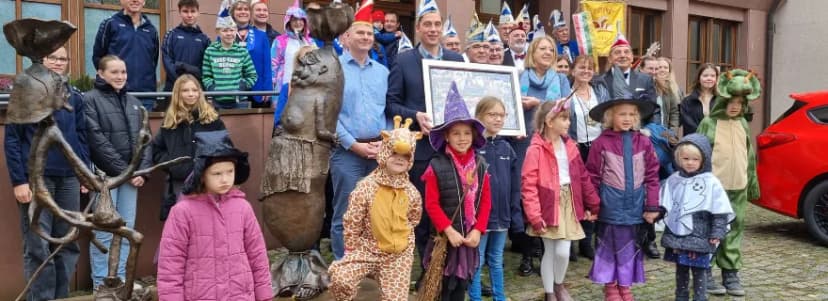 Gruppenfoto mit kostümierten Kindern (Hexe, Giraffe, Drache) und Erwachsenen in Faschingsnarrenkappen vor einem Gebäude mit Bronzefiguren, zwei Männer halten ei