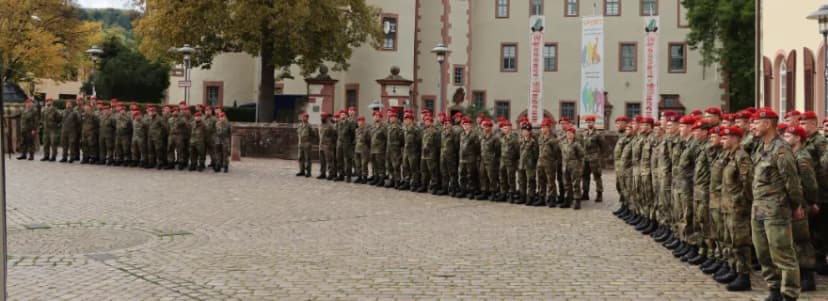 Soldaten der Bundeswehr mit roten Baretten stehen in Reihen auf einem Kopfsteinpflasterplatz vor einem historischen Gebäude mit Bannern.