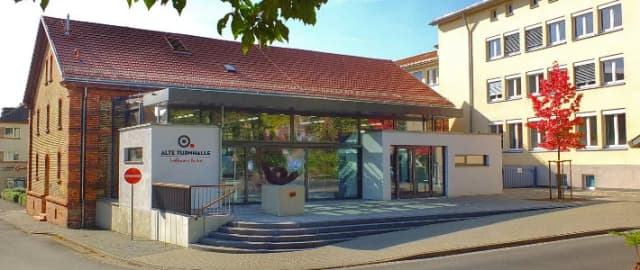 Gebäude der Alten Turnhalle mit Glasfassade und Eingangstreppe, daneben ein Backsteinhaus und ein Wohnhaus mit rot gefärbtem Baum.