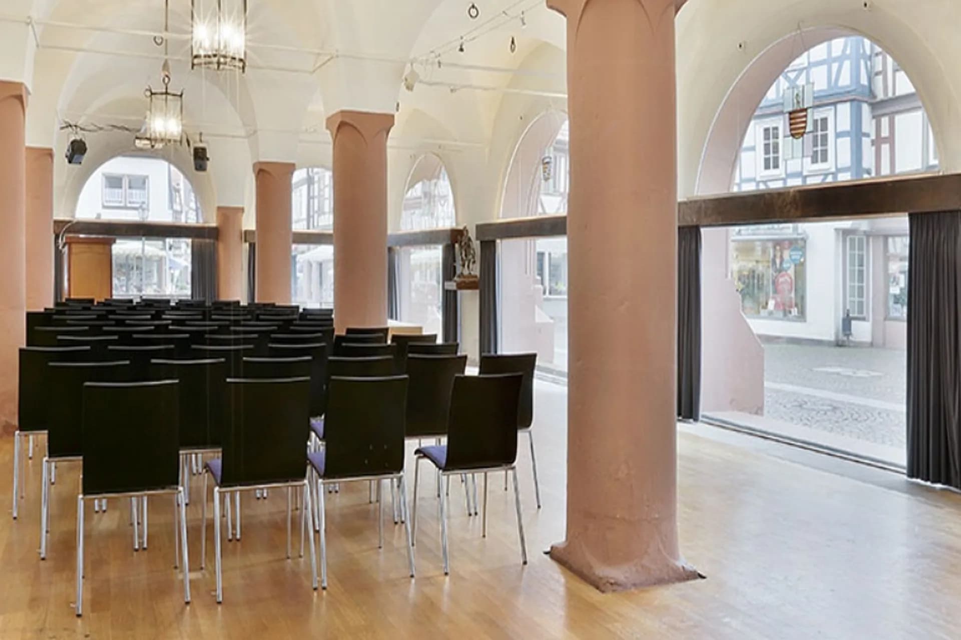 Historische Halle mit gewölbter Decke, Sandsteinsäulen und Stuhlreihen, Bogenfenster mit Blick auf Fachwerkhäuser.