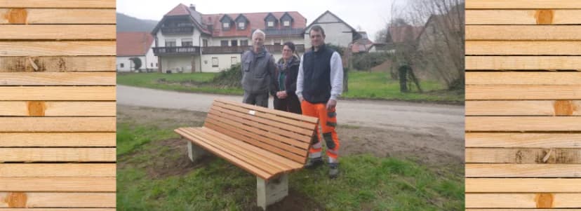 Drei Personen stehen bei einer neu aufgestellten Holzbank auf einer Grünfläche vor einem Wohnhaus