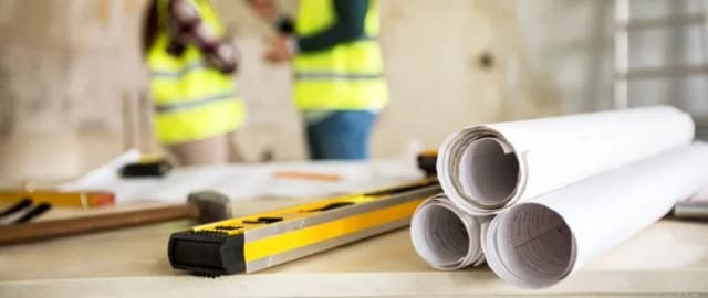 Wasserwaage und aufgerollte Baupläne auf einem Tisch, im Hintergrund Personen in Warnwesten auf einer Baustelle.