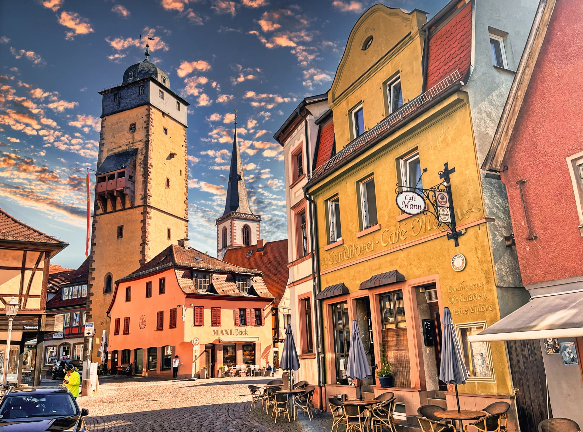 Historischer Stadtturm mit Kirchturm im Hintergrund, umgeben von bunten Fachwerk- und Bürgerhäusern mit Café und Straßencafé-Tischen