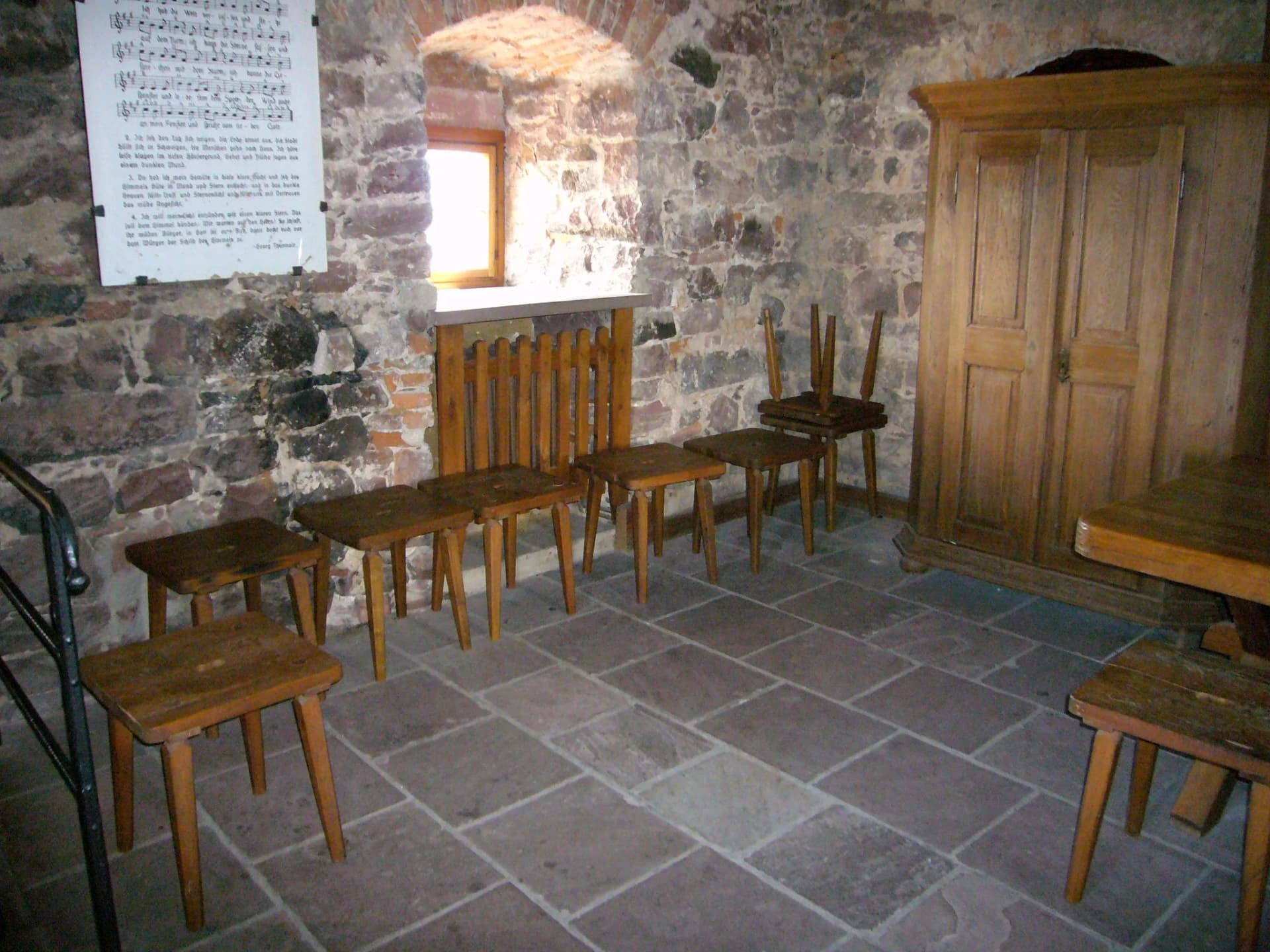 Raum mit Steinmauern im Bayersturm, Holzhocker, Holzschrank und Liedblatt an der Wand