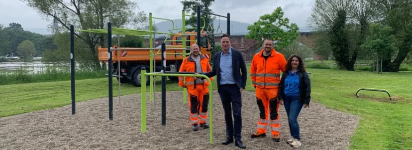 Vier Personen stehen vor einer neuen Calisthenics-Anlage im Park, im Hintergrund ein Arbeitsfahrzeug und der Fluss.