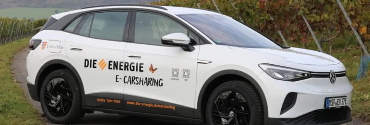 Weißer Volkswagen mit Beschriftung „Die Energie E-Carsharing“ steht auf einem Weg zwischen Weinreben.
