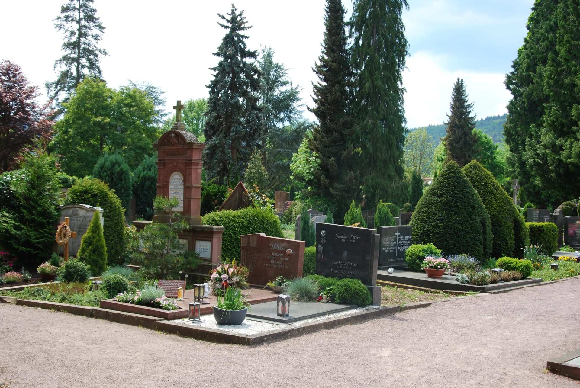 Friedhof mit Grabsteinen aus Sandstein und Granit, umgeben von Bäumen, Hecken und Blumenschmuck.