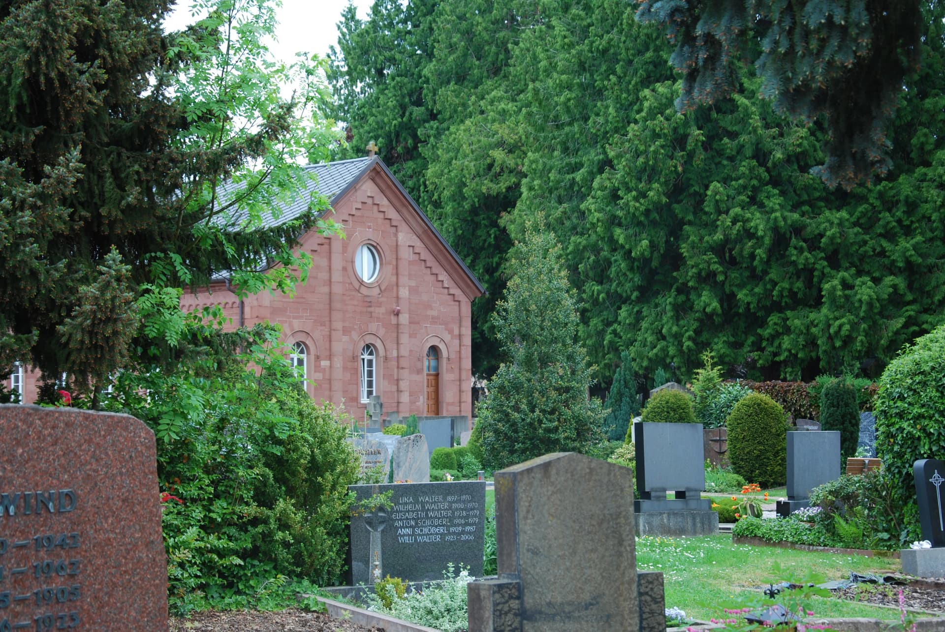 Friedhof mit Grabsteinen, Bäumen und einer roten Sandsteinkapelle im Hintergrund.