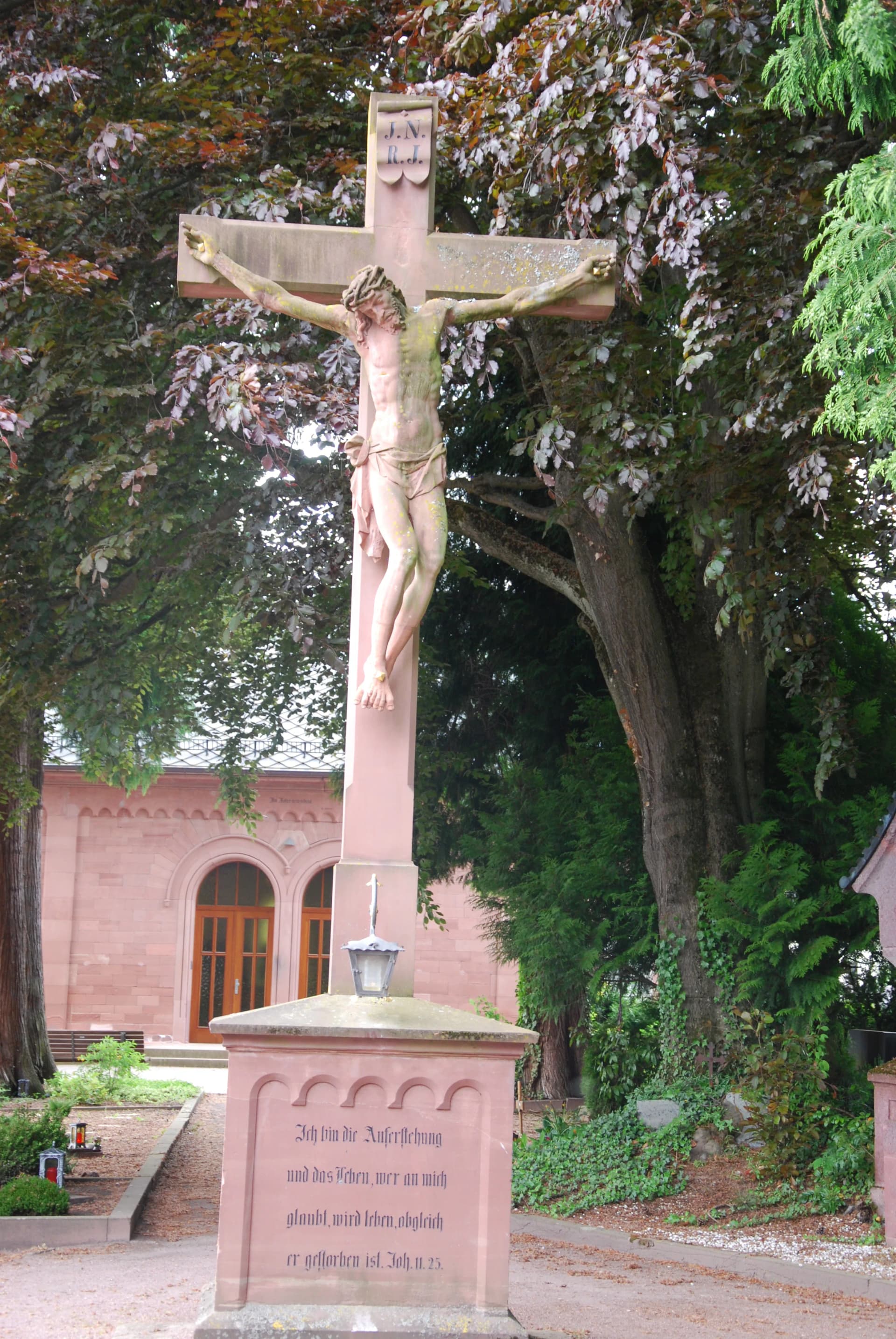 Steinernes Kreuz mit Christusfigur und Bibelinschrift auf einem Friedhof, im Hintergrund eine Kirche