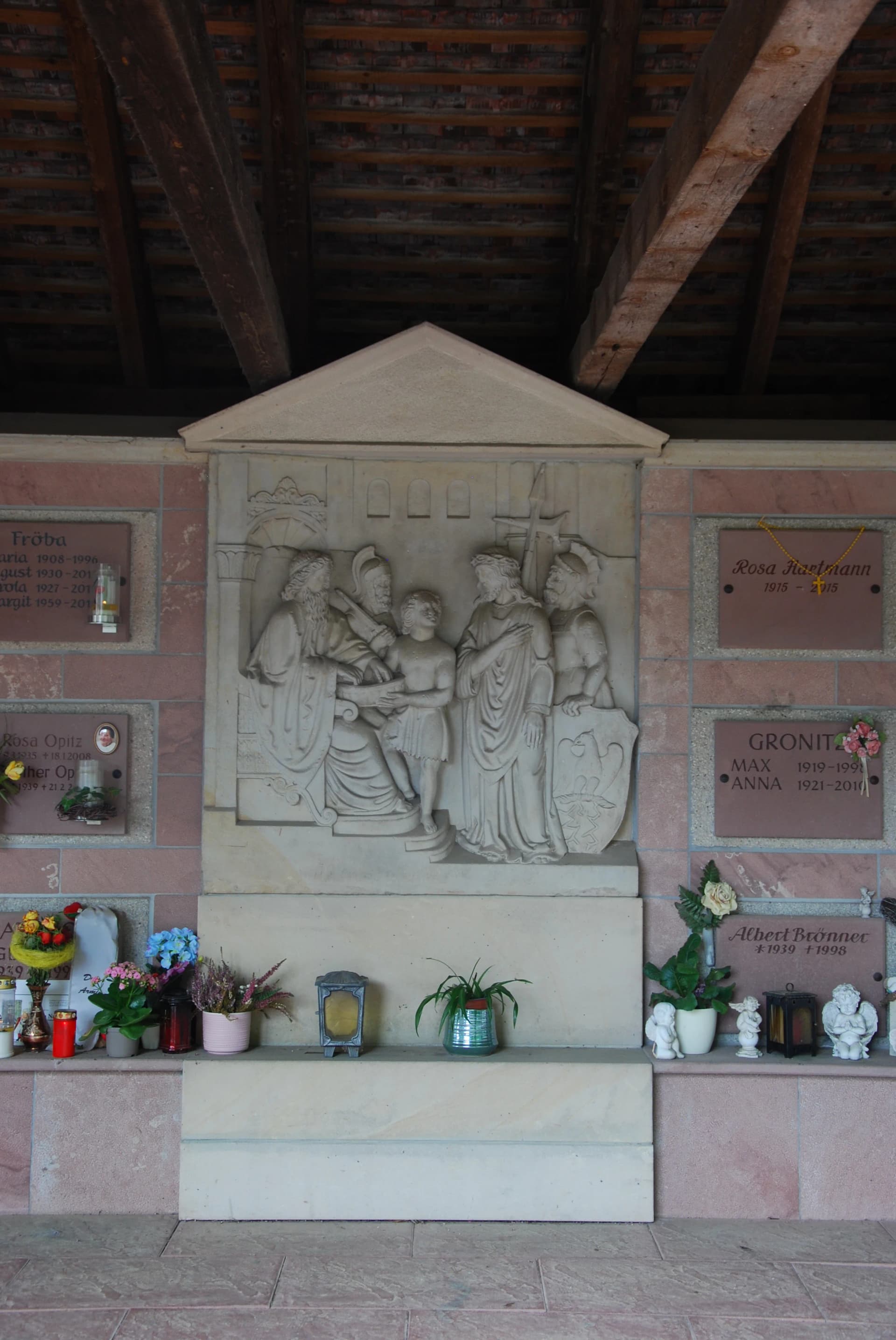 Grabwand mit steinernem Relief religiöser Figuren, umgeben von Namensplatten, Grablichtern und Blumen