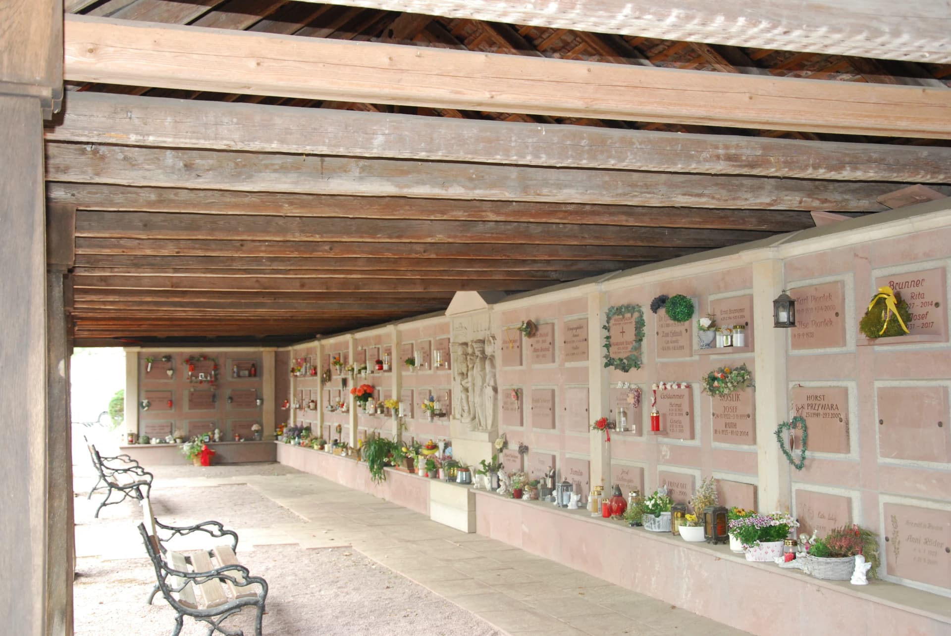 Überdachte Urnenwand auf einem Friedhof mit zahlreichen Gedenktafeln, geschmückt mit Blumen und Kerzen, davor Sitzbänke.