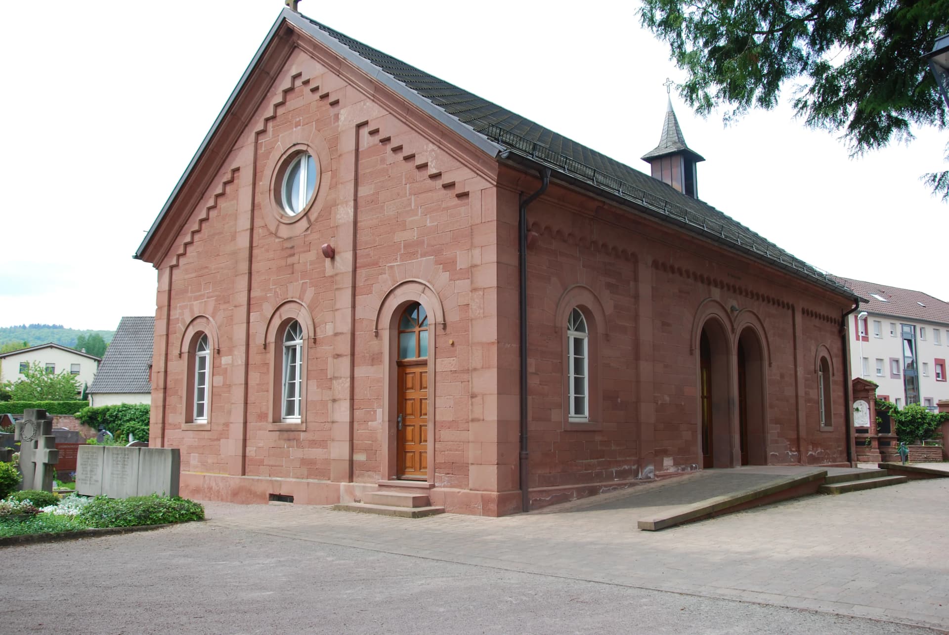 Kapelle aus rotem Sandstein mit rundem Fenster, Dachreiter und Kreuz auf dem Friedhof
