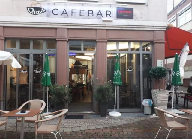 Eingang der Dani Cafebar mit Außenbestuhlung und geschlossenen Sonnenschirmen auf gepflastertem Platz