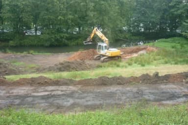 Bagger führt Erdarbeiten an einem Gewässerufer mit aufgeschütteten Erdhaufen durch, im Hintergrund Bäume