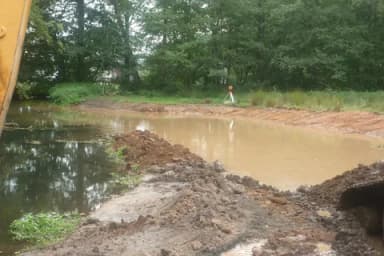 Ausgehobenes Gewässerbecken mit trübem Wasser und Erdaushub, umgeben von Bäumen und Wiese