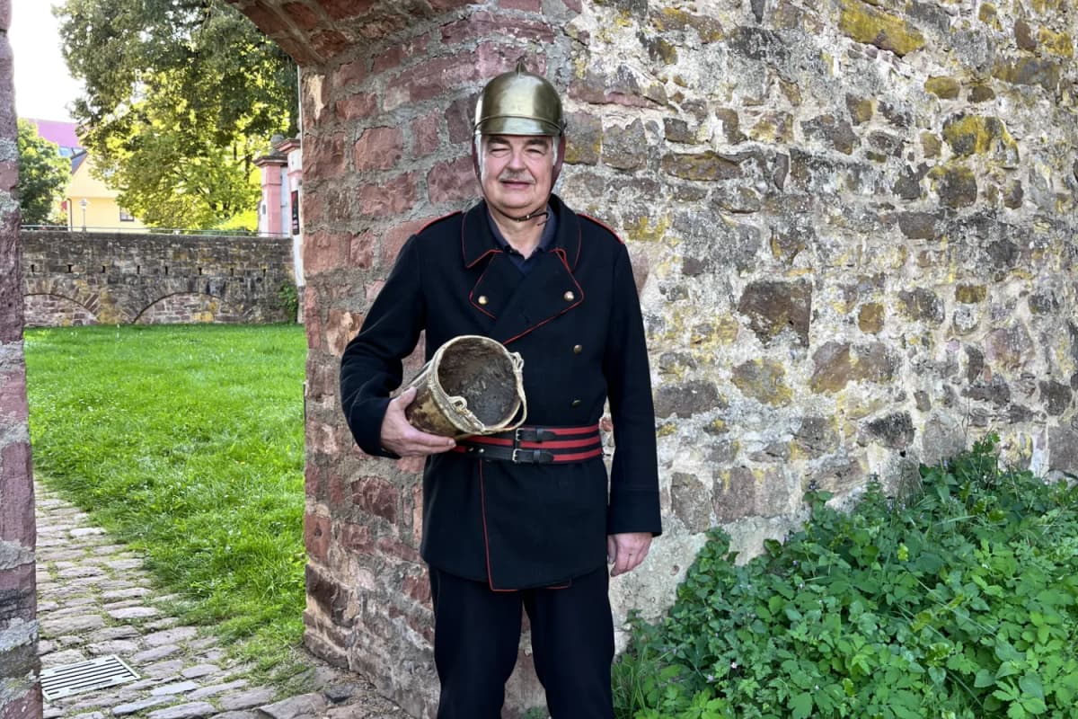 Ein Mann in historischer Feuerwehruniform und mit Messinghelm hält einen alten Löscheimer vor der alten Stadtmauer in Lohr am Main.