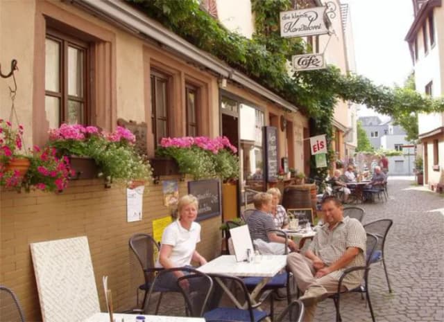Gäste sitzen an Tischen vor einem Café mit Blumenkästen in einer engen Altstadtgasse.