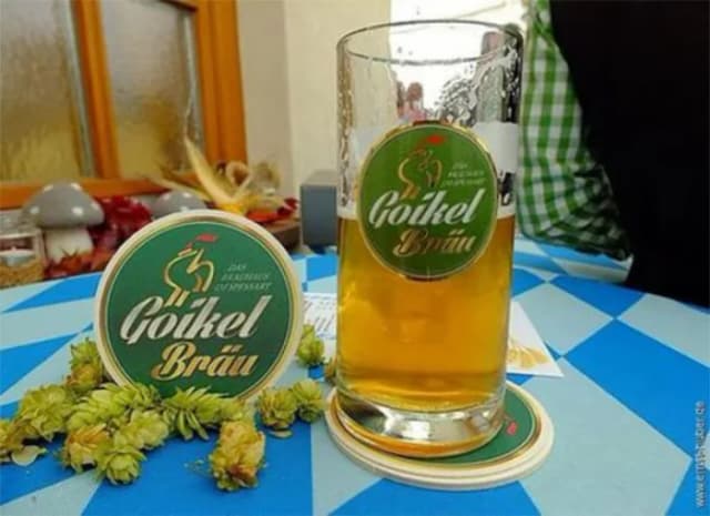 Bierglas mit Goikel Bräu neben Bierdeckel und Hopfenblüten auf blau-weiß kariertem Tisch