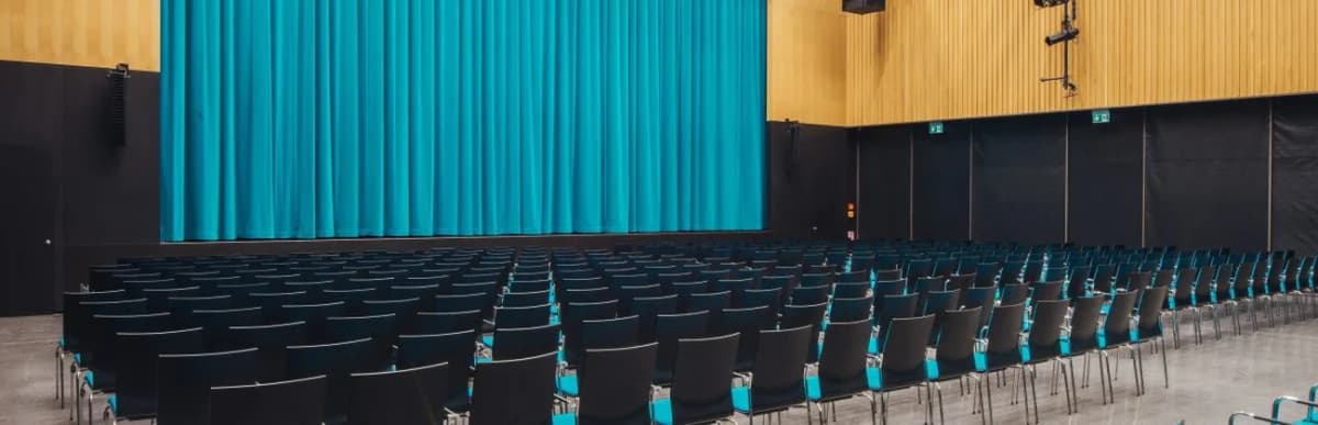 Leerer Saal mit Stuhlreihen und türkisfarbenem Bühnenvorhang