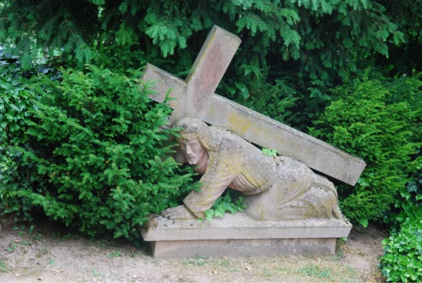 Steinfigur eines Mannes, der kniend ein großes Kreuz trägt, umgeben von grünen Sträuchern auf einem Friedhof