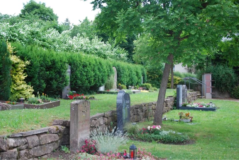 Friedhof mit Grabsteinen, Blumenbeeten und Hecken zwischen grünen Rasenflächen und Bäumen