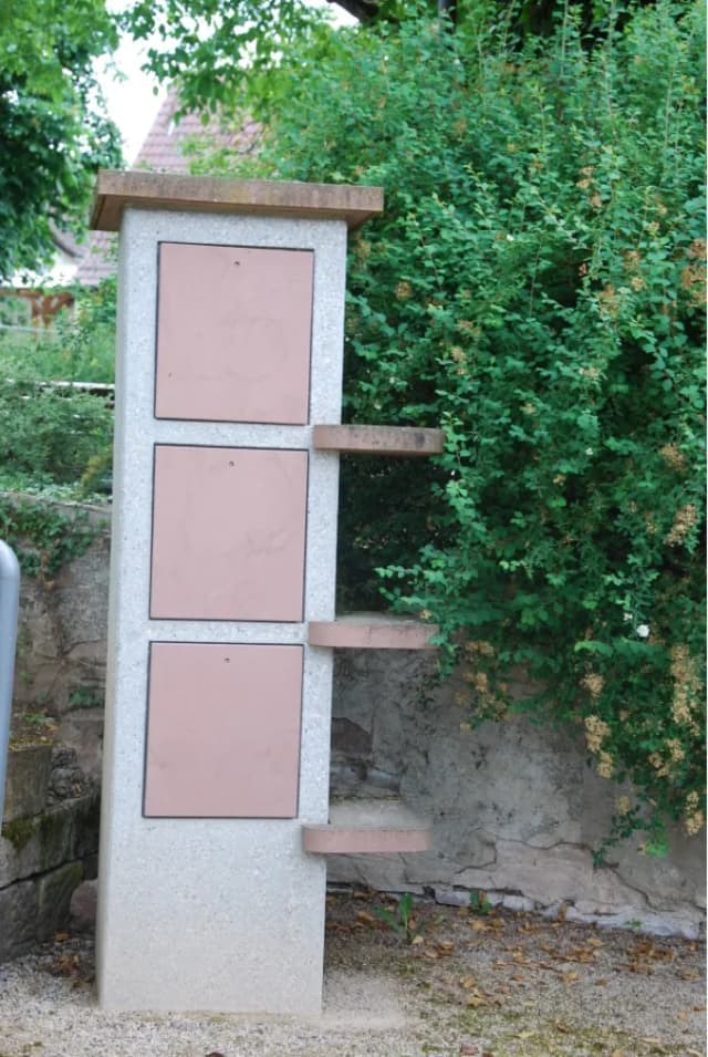 Steinsäule mit drei quadratischen Klappen und Ablageflächen vor einer Hecke und Mauer