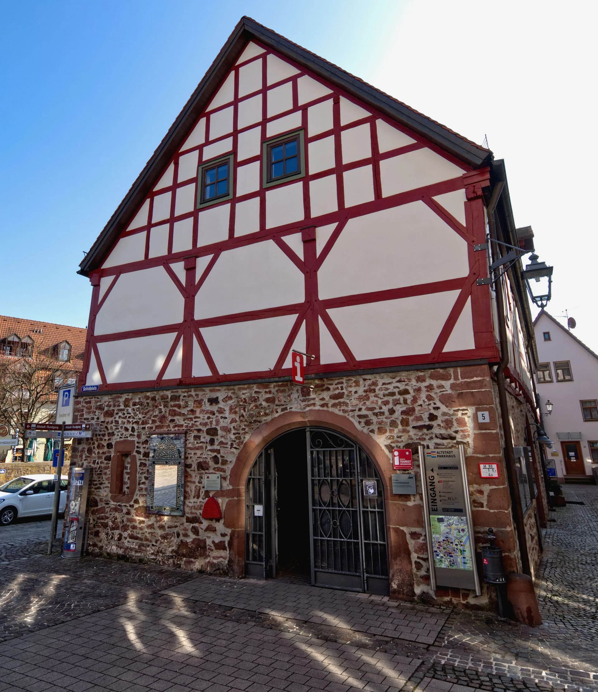 Fachwerkgebäude mit rot-weißer Fassade und steinernem Erdgeschoss, in dem die Touristinformation Lohr untergebracht ist. Darauf weist auch ein Schild hin.
