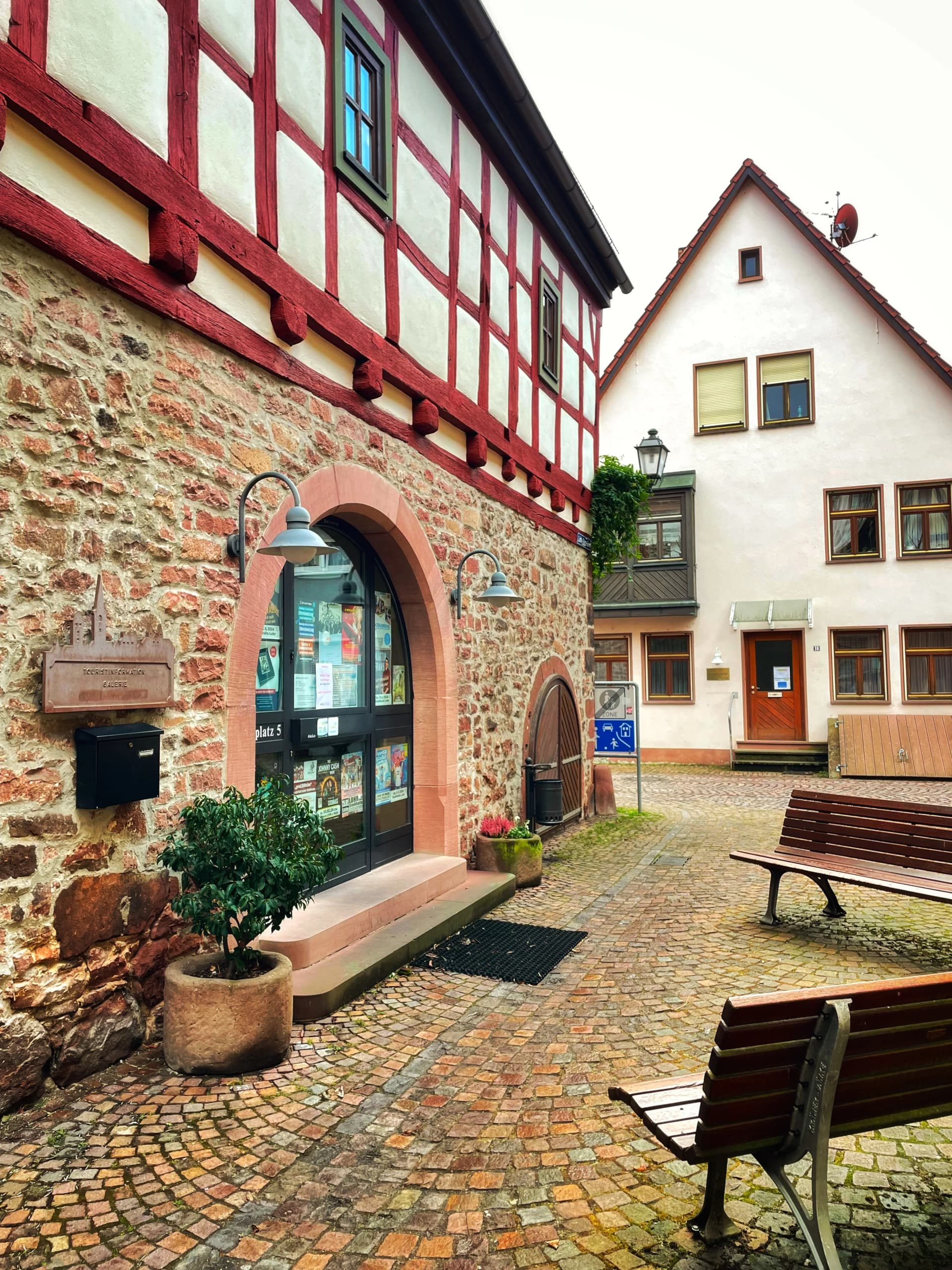 Fachwerkhaus mit Steinsockel und Rundbogeneingang an gepflasterter Gasse, im Hintergrund weitere historische Häuser und Sitzbänke.