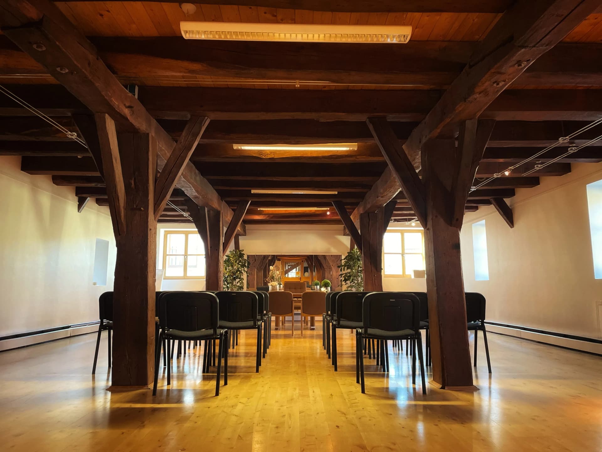 Historischer Veranstaltungssaal mit sichtbarem Holzbalkenwerk, Stuhlreihen und Blick auf gedeckte Tische im Hintergrund.