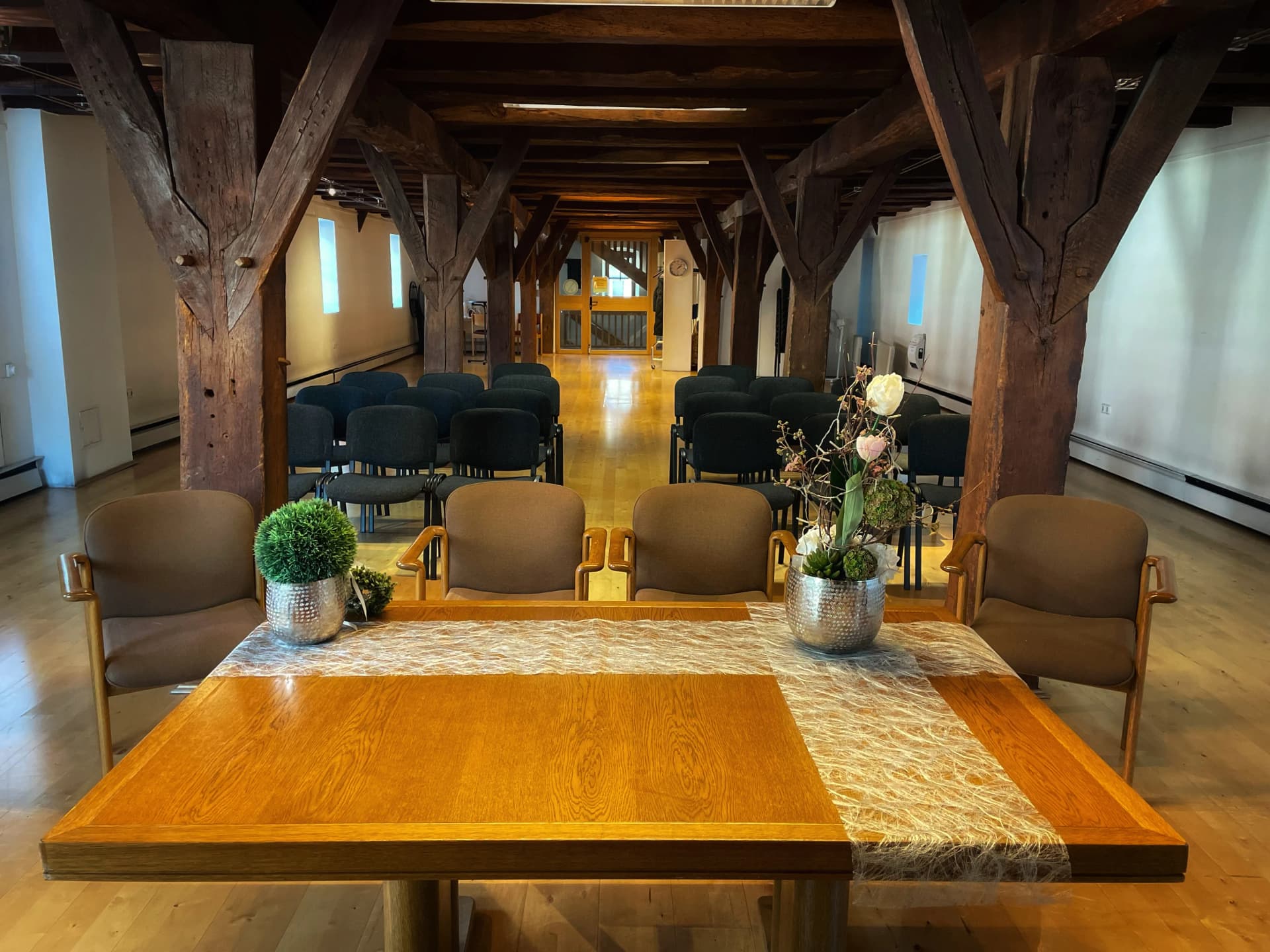 Historische Scheune mit Holzbalken, aufgestellten Stuhlreihen und einem Tisch mit Blumendekoration im Vordergrund.