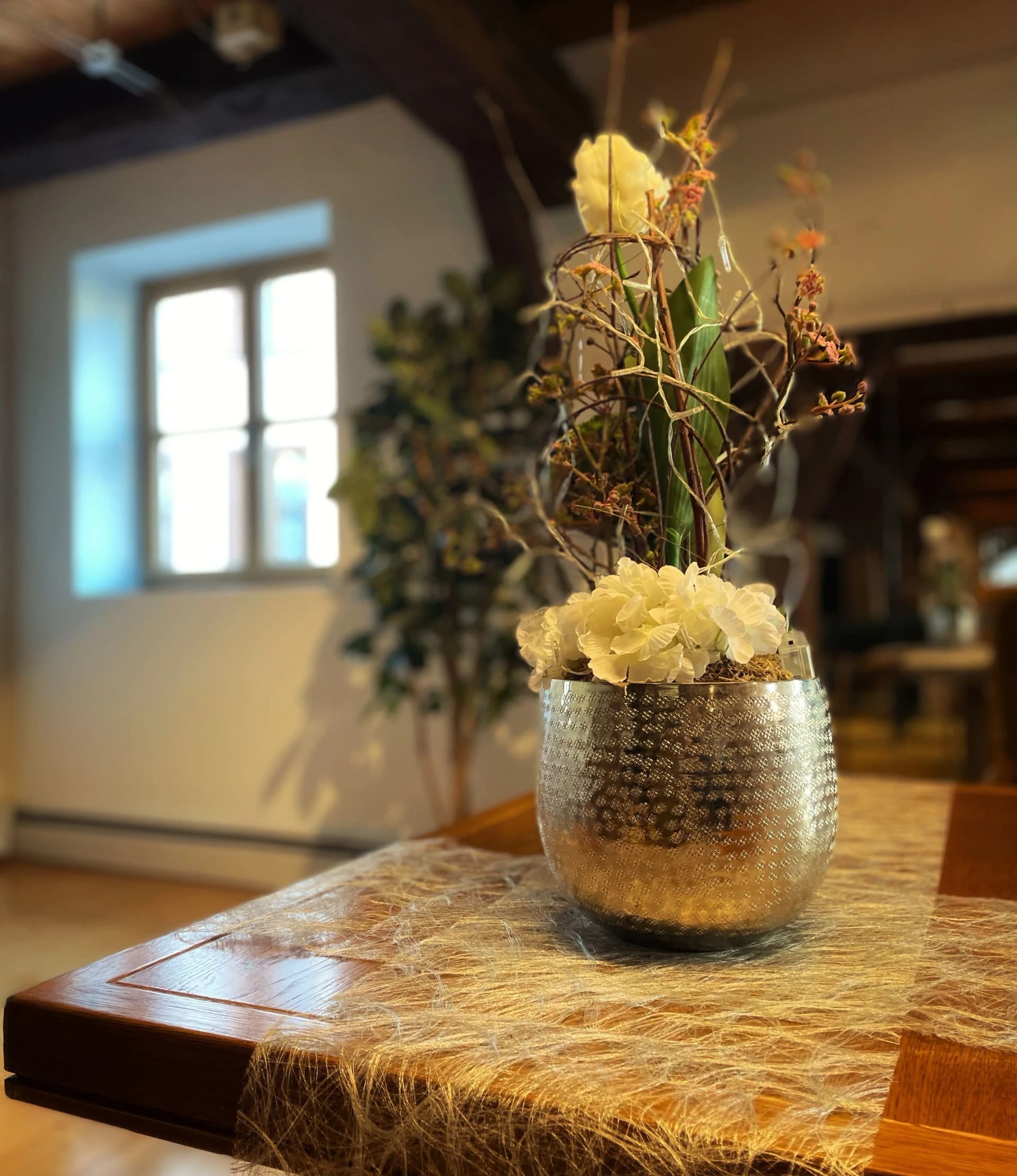Blumengesteck mit weißen Blüten in silberner Metallvase auf einem Holztisch in einem Raum mit Holzbalken