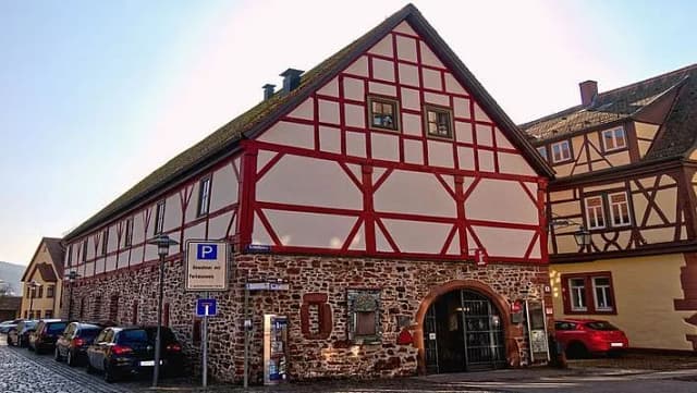 Fachwerkgebäude mit rotem Fachwerk auf steinernem Sockel, davor parkende Autos und ein Parkschild