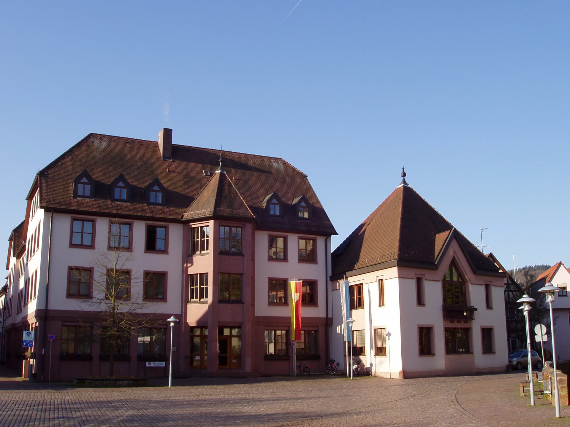 Rathausgebäude mit Fahnenmasten an einem gepflasterten Platz unter blauem Himmel