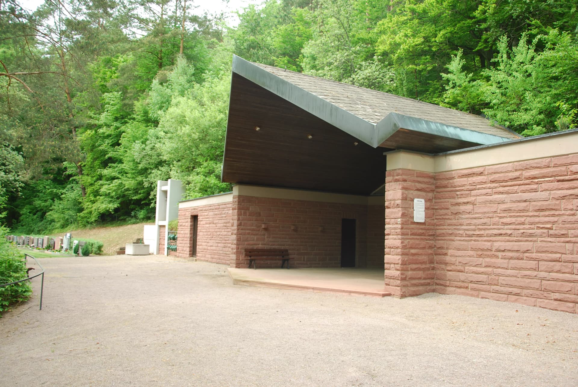 Friedhofsgebäude mit Sandsteinmauern und geschwungenem Dach vor einem Wald, davor ein Kiesplatz