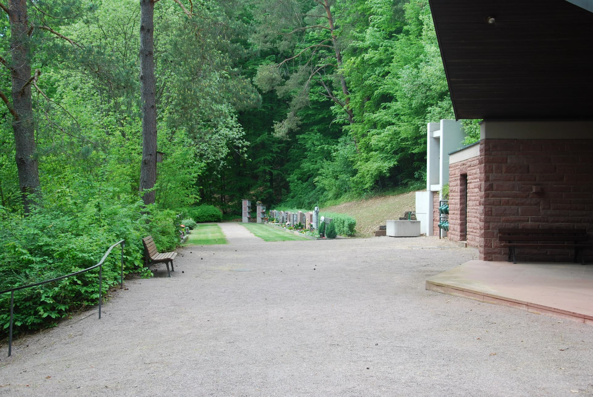 Kiesweg an einem Friedhofsgebäude mit Blick auf Grabreihen am Waldrand