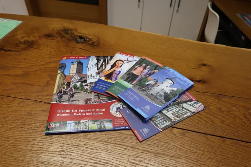 Ein Katalog und mehrere Flyer der Touristinformation Lohr am Main liegen auf einem Holztresen. Zu sehen sind zum Beispiel der Urlaubkatalog oder Flyer zu den Themen Wandern, Radfahren, Altstadt und Schneewittchen.