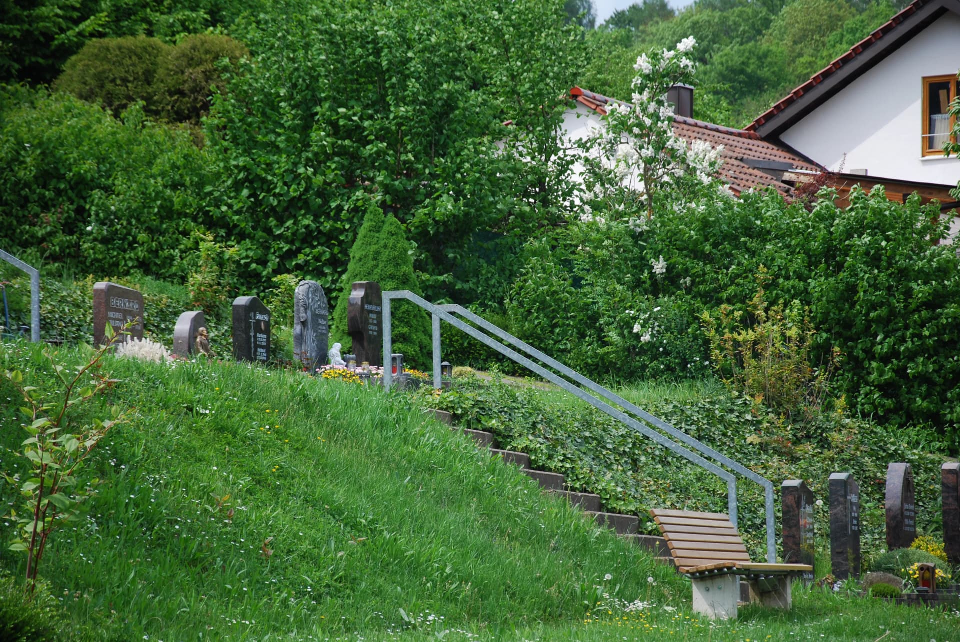 Friedhofsgelände an einem grünen Hang mit Treppe, Geländer, Grabsteinen und einer Sitzbank, im Hintergrund ein Wohnhaus.