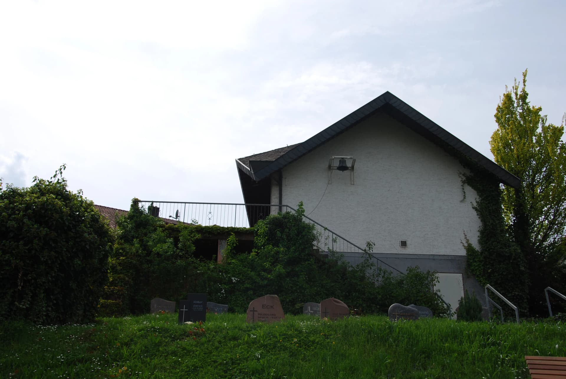 Weißes Gebäude mit Glocke am Giebel und Freitreppe, davor ein Friedhof mit Grabsteinen