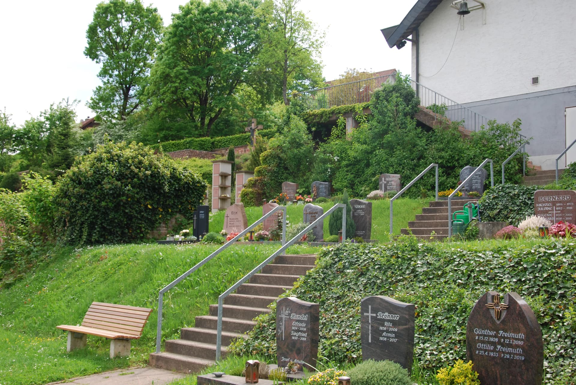 Terrassenförmig angelegter Friedhof an einem Hang mit Treppen, Grabsteinen, Grün und einer Sitzbank.