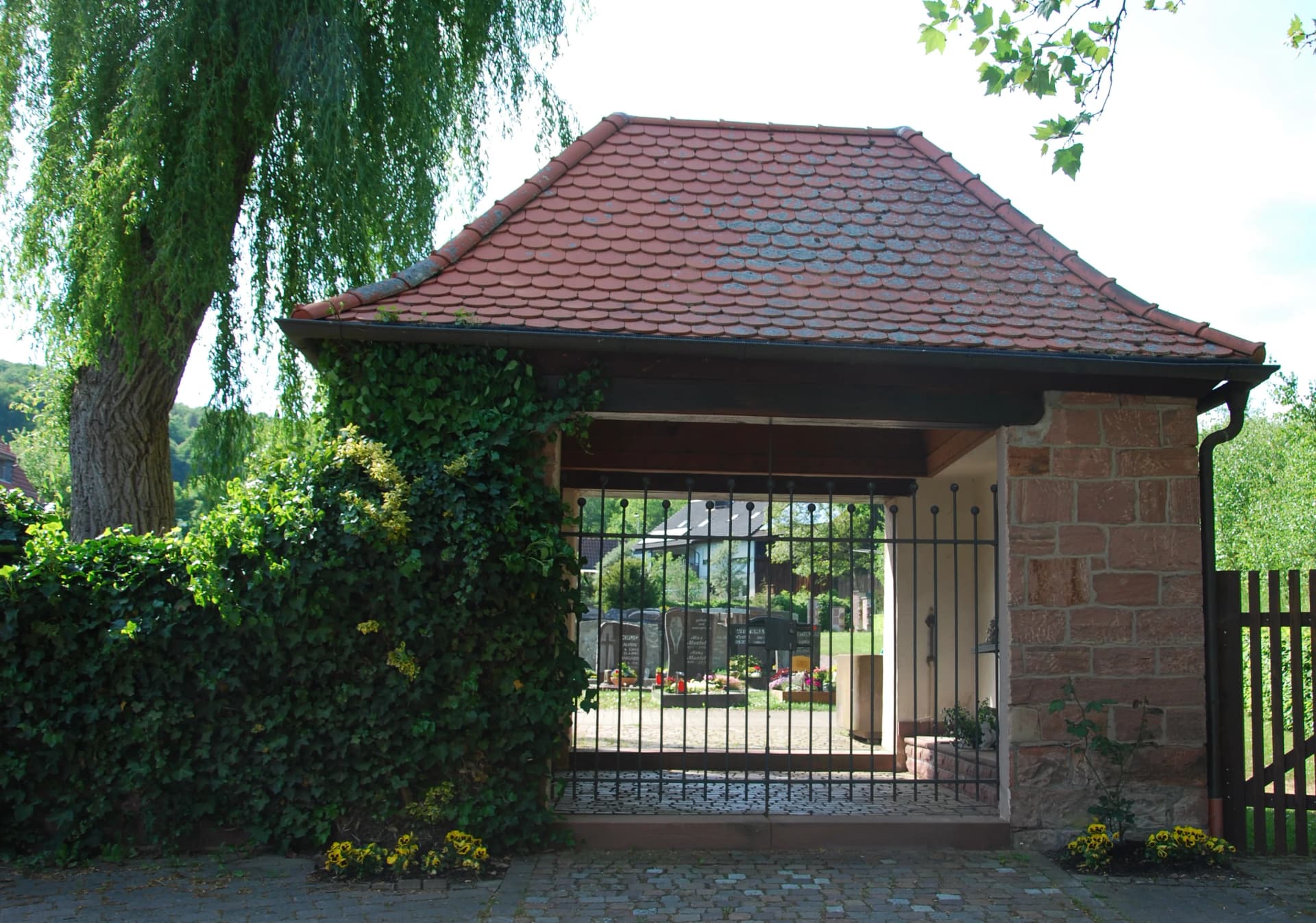 Überdachtes Friedhofstor mit Ziegeldach und schmiedeeisernem Gitter, umgeben von Efeu, Blick auf Grabreihen dahinter