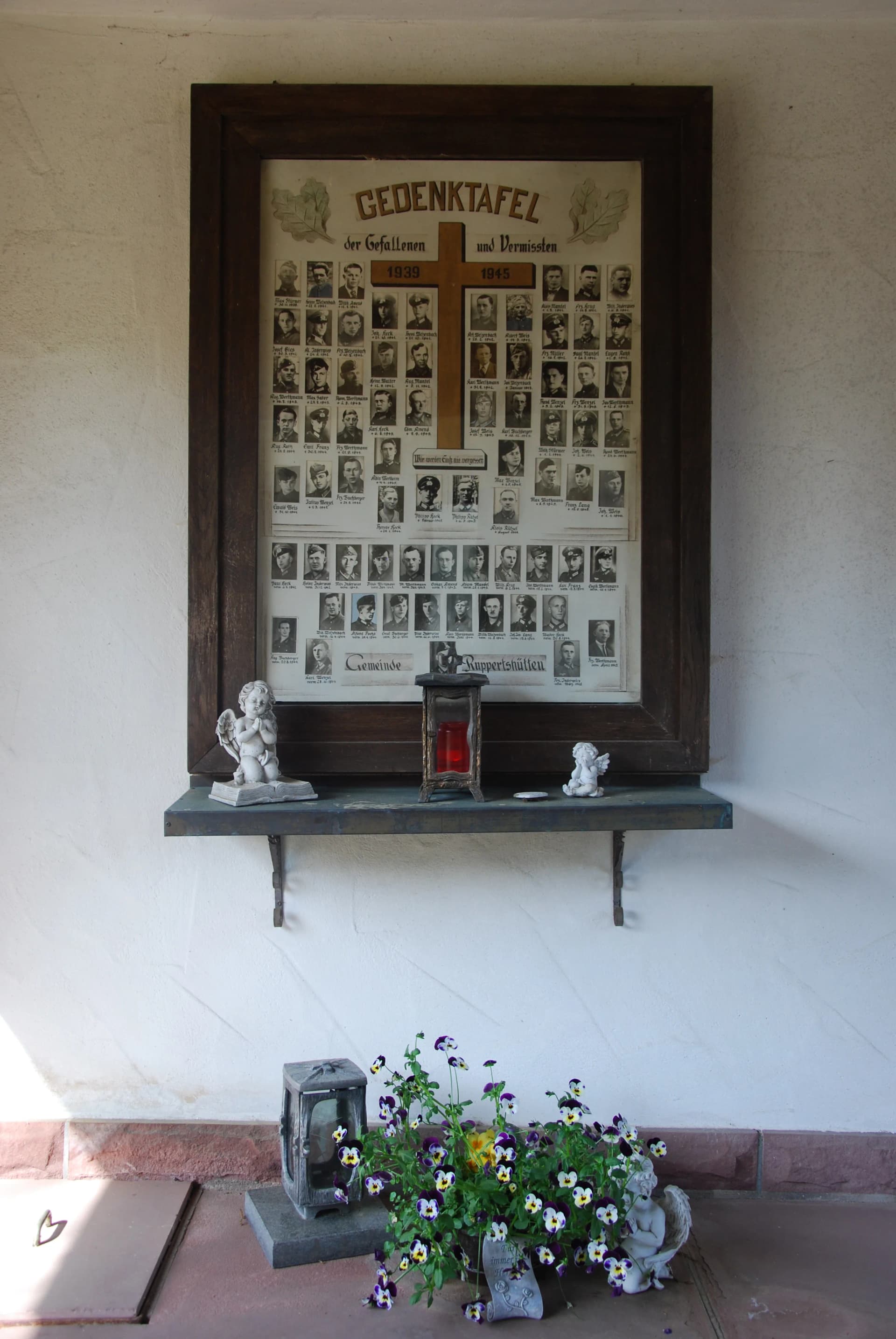 Gedenktafel für Gefallene und Vermisste 1939–1945 mit Porträtfotos in einem Holzrahmen, daneben Blumenschmuck an einer Hauswand