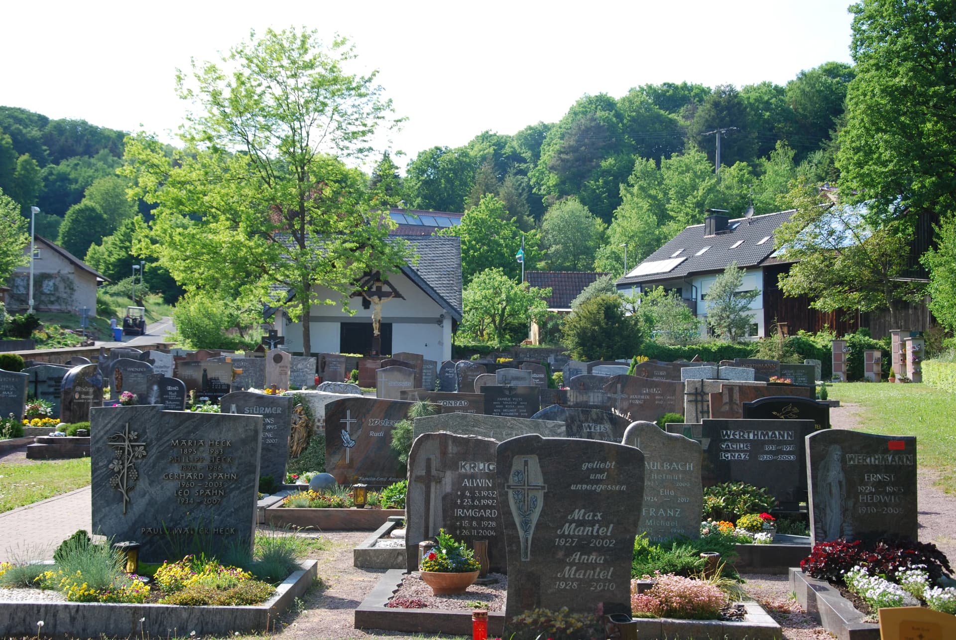 Friedhof mit zahlreichen Grabsteinen, im Hintergrund ein Kreuz, Häuser und bewaldeter Hang