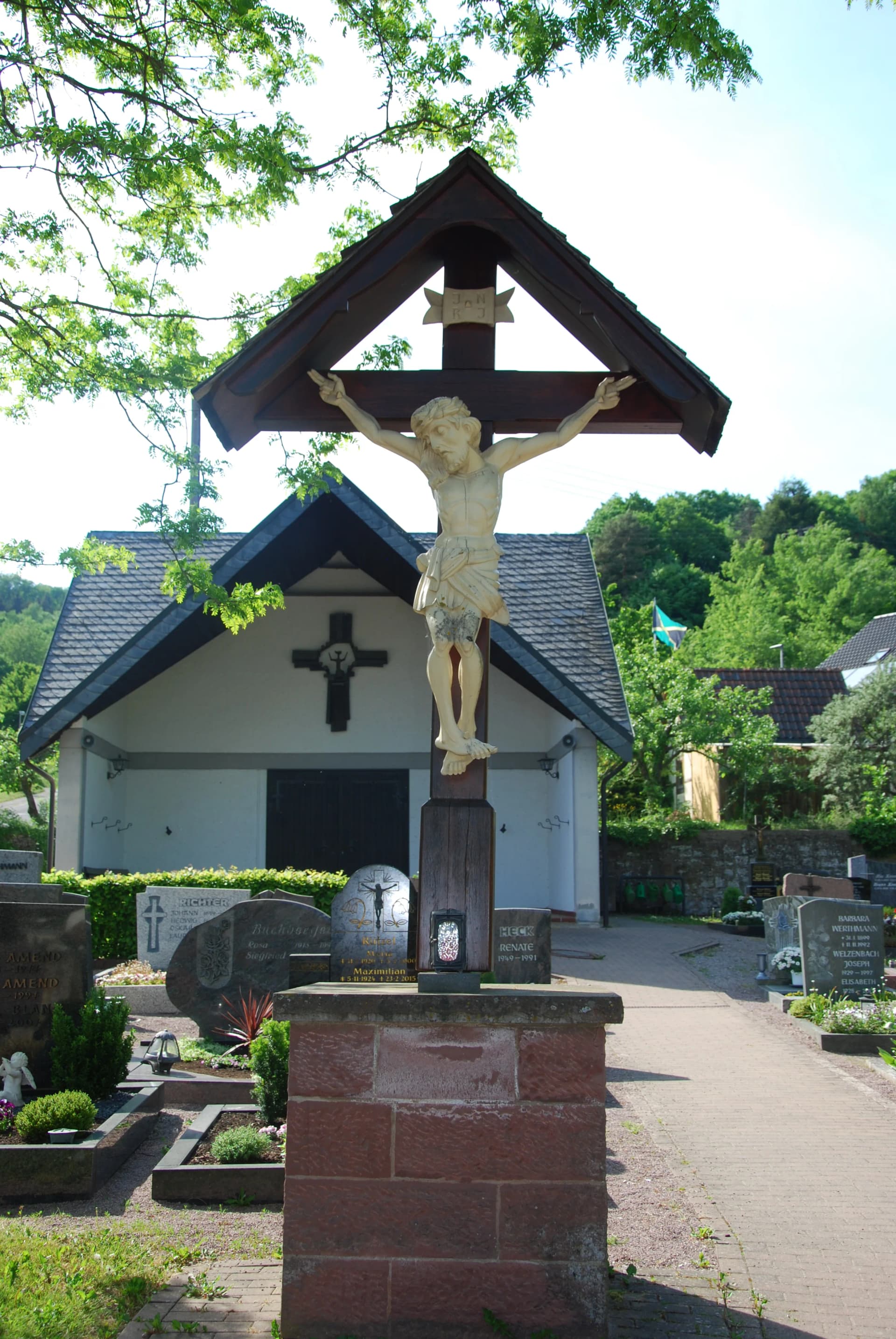 Kruzifix mit überdachtem Vordach auf einem Friedhof, umgeben von Grabsteinen und Bäumen